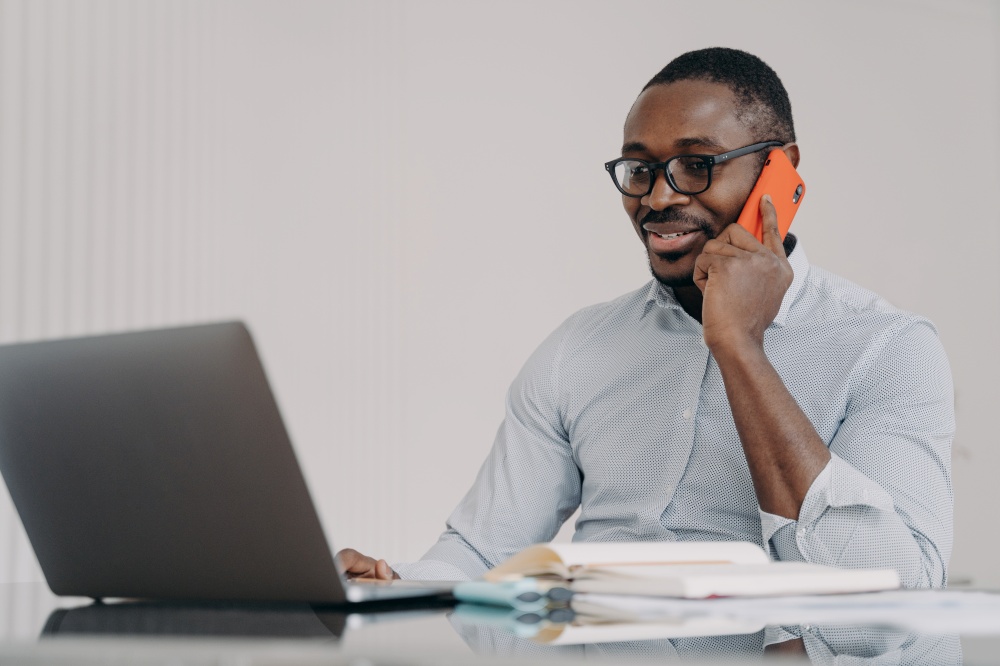 African american businessman talks on phone at laptop in office. Black male manager wearing glasses consulting client, having pleasant conversation with colleague, discussing solving problem distantly. African american businessman talk on phone at laptop, consulting client, discussing business project