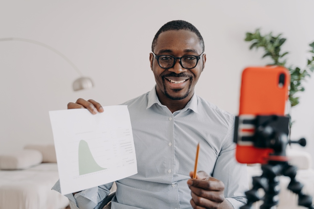 Afro freelancer has online brainstorm. Positive african american man shows data chart. Businessman is speaking at online conference from home on quarantine. Distant study, remote education.. Afro freelancer has online brainstorm. Positive man shows data chart speaking at online conference.