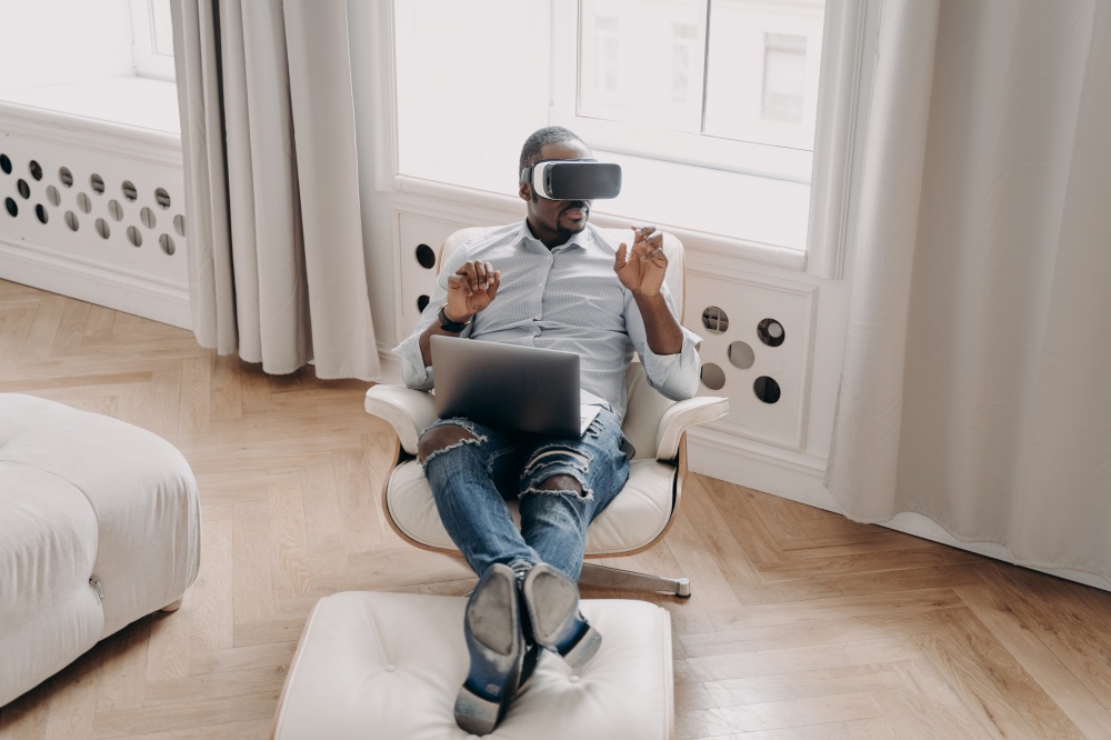 African american man in VR glasses working in augmented reality, sitting in armchair with laptop at home, pointing at virtual reality object, interacting with cyberspace, using modern high-tech device. African american businessman in VR glasses works in virtual reality, sitting in armchair with laptop