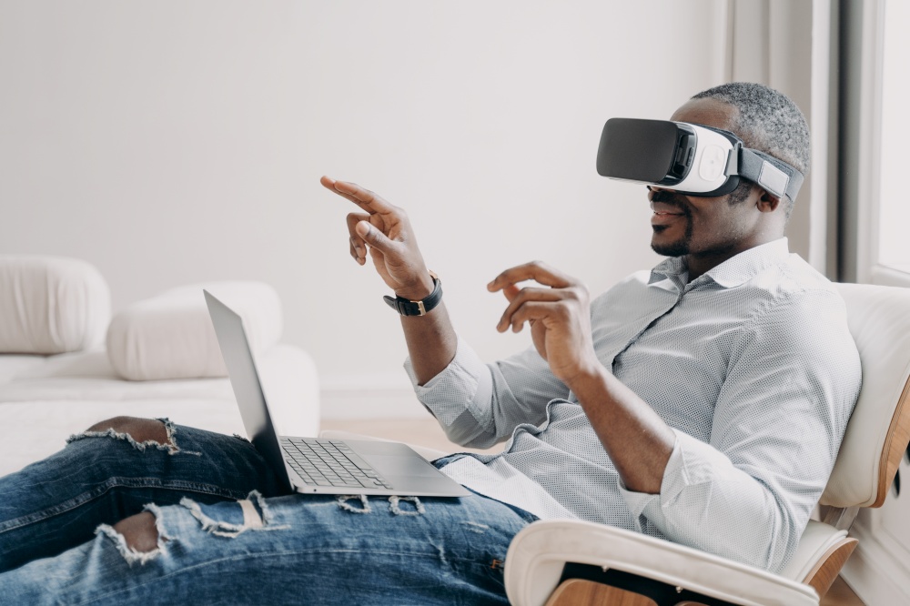 Relaxed businessman clicks virtual buttons. Gestures of african american man in vr glasses. Guy is working at home. Distant work and futuristic gaming. Modern digital technology for business.. Relaxed businessman clicks virtual buttons. Gestures of african american man in vr glasses.