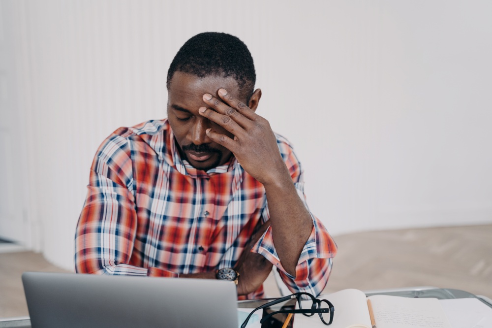 Sad tired online teacher or businessman. Afro man has online conference on laptop or working on project late. Tutor is exhausted from work. Remote study, distance learning. Deadline concept.. Sad tired online teacher or businessman. Remote study or work, distance learning. Deadline concept.
