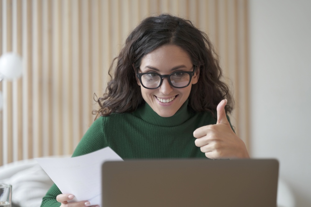 Happy young Italian female employee in glasses talks online on laptop with colleagues or business partner, showing thumb up, cheerful Spanish woman freelancer making video call while working remotely. Happy young Italian female employee talks online on laptop with colleagues and showing thumb up