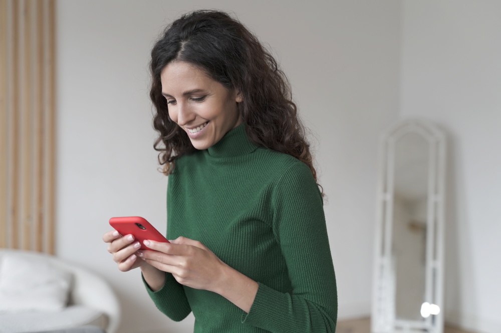 Young happy smiling Spanish woman using mobile phone and chatting with friends, texting sms or checking email while relaxing at home, pleased Italian female holding smartphone enjoying online shopping. Young happy Spanish woman using mobile phone and chatting with friends, texting sms on smartphone