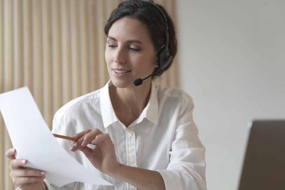 Portrait of positive focused Spanish business woman in headset sits at desk having video conference online with partner, holds accounting report in hand points to calculations in pencil. Positive Spanish businesswoman in headset sits at desk having video conference online with partners
