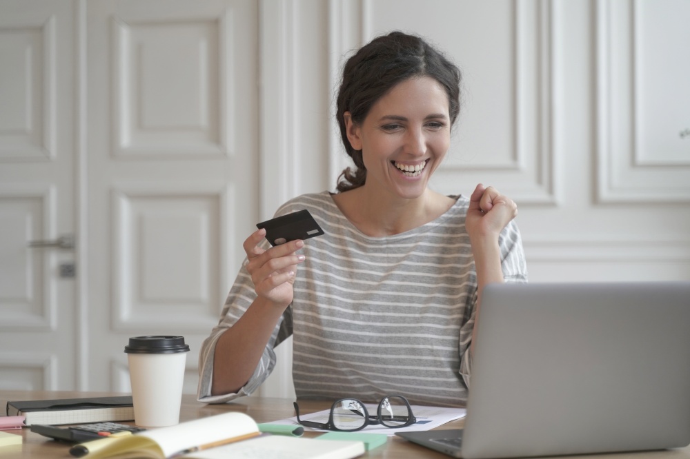 Young happy Spanish woman holding credit card and feeling excited while doing online shopping on laptop at home, looking at computer screen and purchasing goods or services in internet store. Young happy Spanish woman hold credit card and feeling excited while doing online shopping on laptop