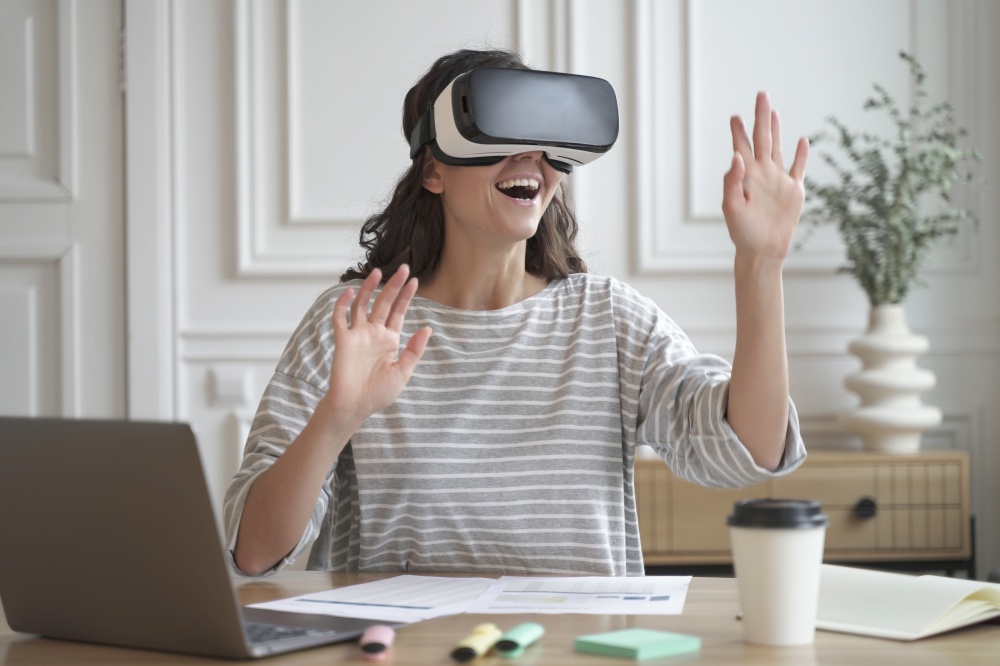 Photo of excited amazed woman wearing VR glasses enjoying amazing virtual reality experience while sitting at office table, making purchases in 3d store. Innovation and technology concept. Excited amazed woman wearing VR glasses enjoying amazing virtual reality experience at work