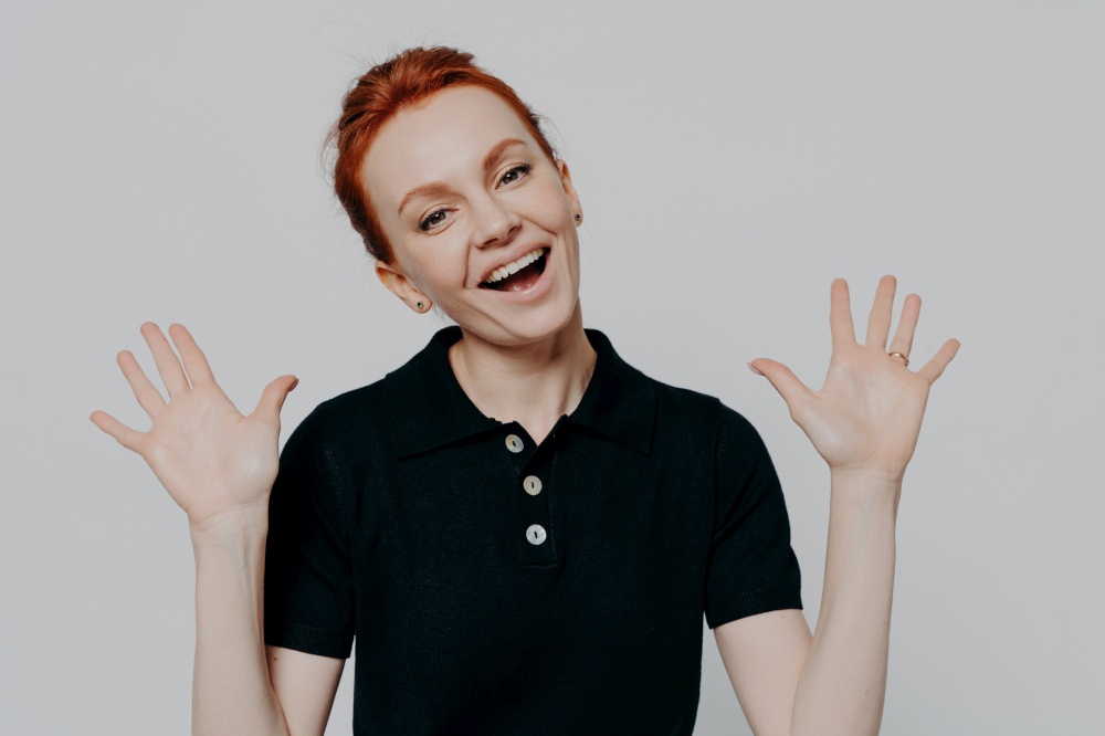 Awesome news. Studio shot of young excited red haired woman raising palms and exclaiming loudly while posing isolated on grey background, keeping mouth opened and dressed in black t-shirt. Young excited red haired woman raising palms and exclaiming loudly, isolated over grey background