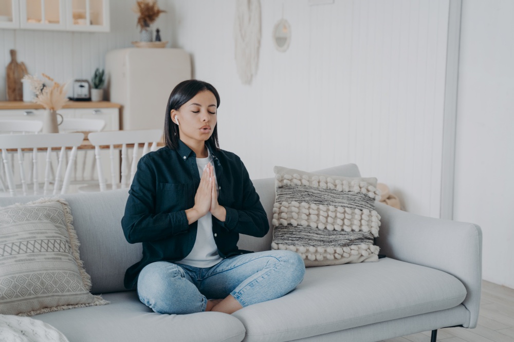Calm young woman deep breathing practicing yoga sitting on couch at home. Female meditates folded hands in namaste gesture in lotus posture on sofa. Wellness, stress relief concept.. Calm woman breathing practicing yoga sitting in lotus pose on couch at home. Wellness, stress relief