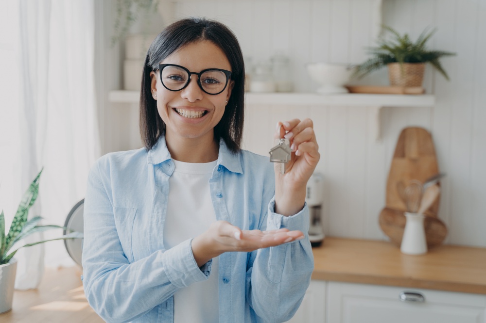 Smiling positive spanish woman is showing house key in her hand. Success and achievement concept. Happy real estate owner. Mortgage loan and investment. Modern scandinavian interior of kitchen.. Smiling positive spanish woman is showing house key in her hand. Happy real estate owner.