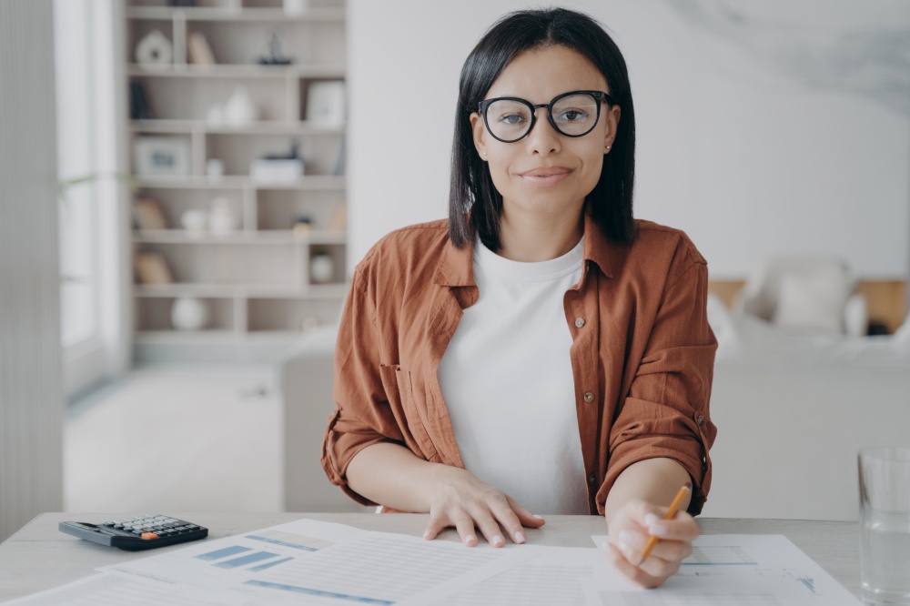 Young european woman in glasses is working remote. Confident manager is doing research at home. Happy business assistant is sitting at the desk and doing paperwork. Strategy planning, data analysis.. Manager doing research at home. Happy business assistant is planning strategy, doing data analysis.