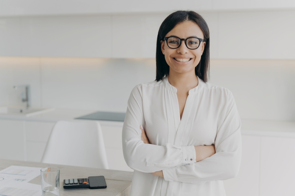Young european executive or ceo in glasses and white blouse is working remote from home. Confident girl, entrepreneur is standing with her arms crossed and smiling. Career and leadership concept.. Young european executive or ceo standing with her arms crossed and smiling. Career and leadership.