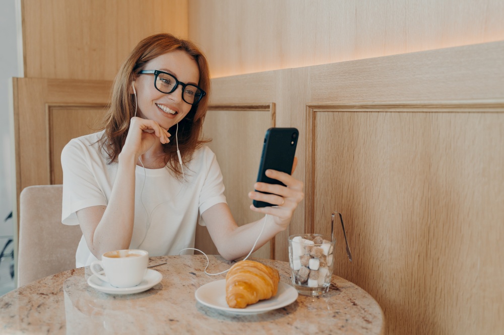 Beautiful redhead woman in earphones sitting in cafe and making video call on mobile phone, talking with friends or followers during video chat on smartphone, blogger enjoying breakfast in coffee shop. Beautiful redhead woman in earphones sitting in cafe and making video call on mobile phone