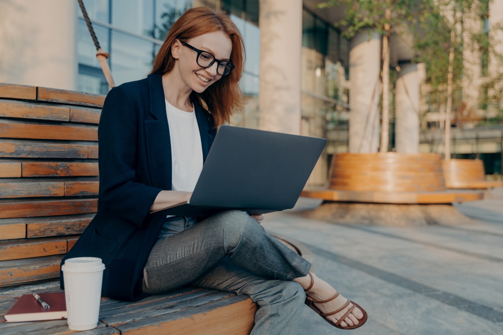 Happy cheerful european ginger woman working remotely on laptop outside, sitting on wooden bench and drinking morning coffee, female in eyeglasses enjoying remote work outdoors, checking e-mail. Happy cheerful european ginger woman working remotely on laptop outside, sitting on wooden bench