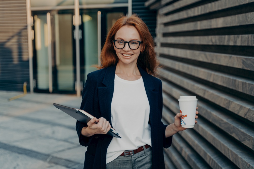 Happy beautiful red-haired business woman in stylish formal outfit with laptop and paper cup of coffee in hands outdoors, smiling at camera, standing outdoors, enjoying lunch break in office. Happy beautiful red-haired businesswoman with laptop and paper cup of coffee in hands outdoors