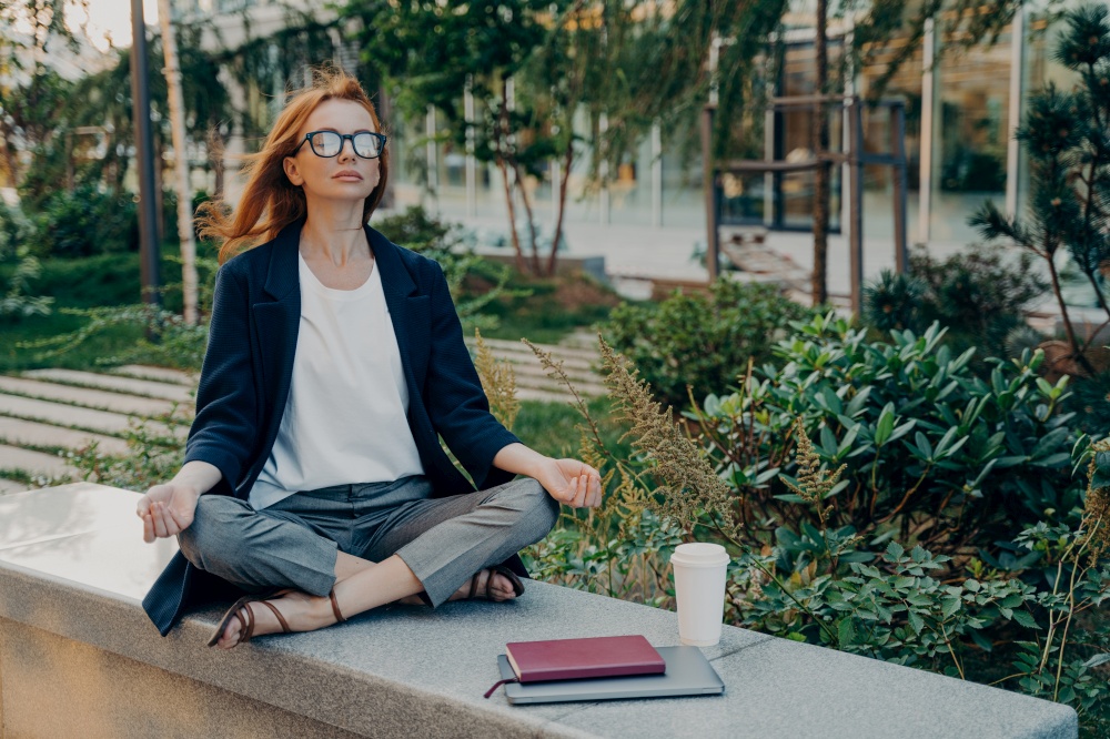 Peaceful redhead business woman dressed formally holding hands in mudra gesture, sitting in lotus pose on concrete bench and meditating with closed eyes, relaxing after remote work on laptop in park. Peaceful business woman holding hands in mudra gesture, sitting in lotus pose and meditating in park