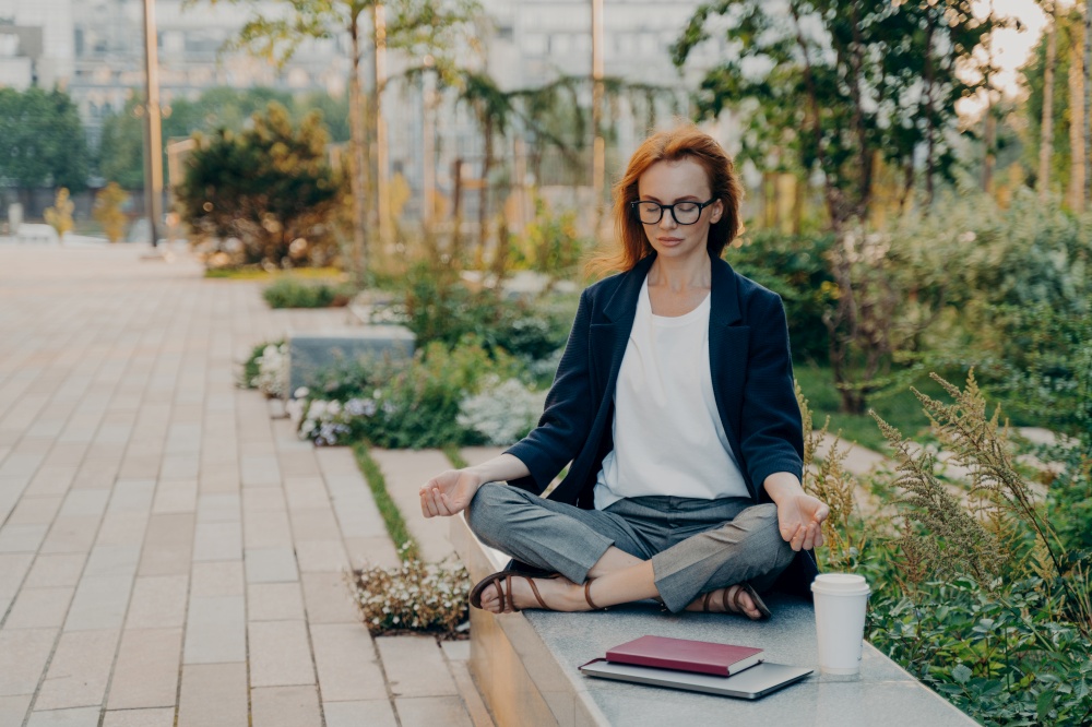 Young relaxed redhead woman meditates in park feels calm sits in lotus pose outdoor wears transparent glasses formal outfit drinks takeaway coffee uses notepad and tablet. Healthy lifestyle meditation. Young relaxed redhead woman meditates in park feels calm sits in lotus pose outdoor