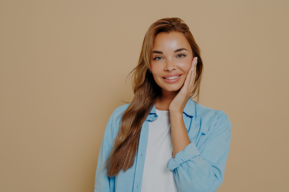 Lovely woman touching her face with clean healthy skin, looking at camera with smile and expressing positiveness. Happy female in casual outfit feeling pleasant while posing against beige background. Lovely woman touching her face with clean healthy skin