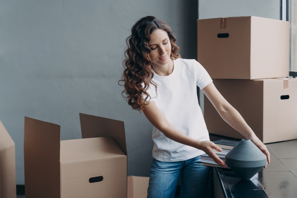 Happy girl unpacking cardboard boxes and taking the vase. Smiling attractive hispanic woman packing boxes to move. Relocation to new apartment. Mortgage and real estate concept.. Happy girl unpacking cardboard boxes. Relocation to new apartment. Mortgage and real estate concept.
