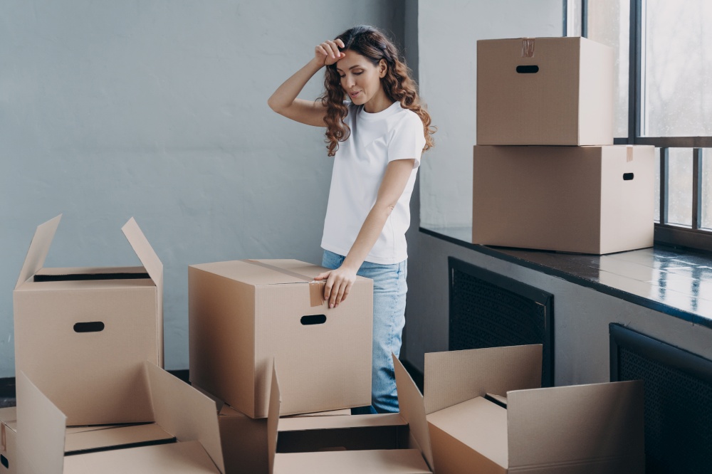 Puzzled woman unpacking cardboard boxes. Tired girl in new apartment. Lady touches forehead with hand, gesture of tired person. Packing things, shipping service and relocation concept.. Tired woman unpacking cardboard boxes. Packing things, shipping service and relocation concept.