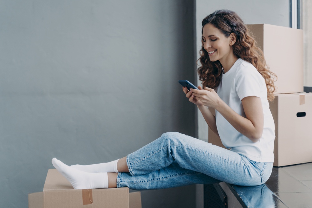 Happy attractive hispanic woman is sitting on the sill with cardboard boxes. Girl is texting on mobile phone. Young lady is unloading boxes. Distance friendship and relocation concept.. Happy attractive hispanic woman is sitting on the sill with boxes and texting on mobile phone.