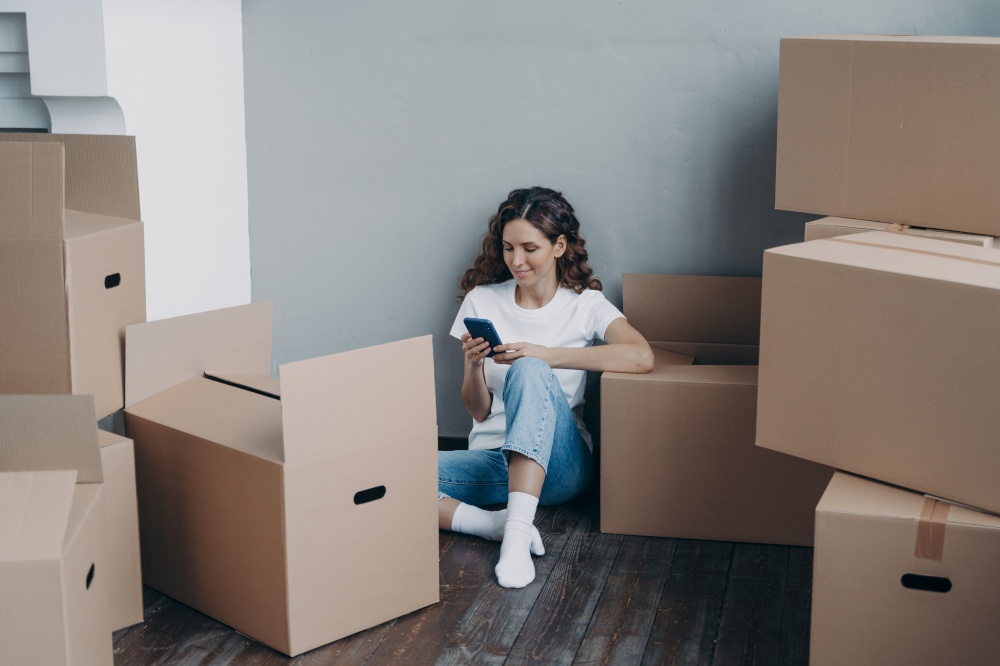 Happy young european woman with smartphone is packing boxes. Girl is chatting on mobile phone and sitting on the floor. Easy moving and shipping service order concept.. Happy young european woman with smartphone is packing boxes. Shipping service order concept.
