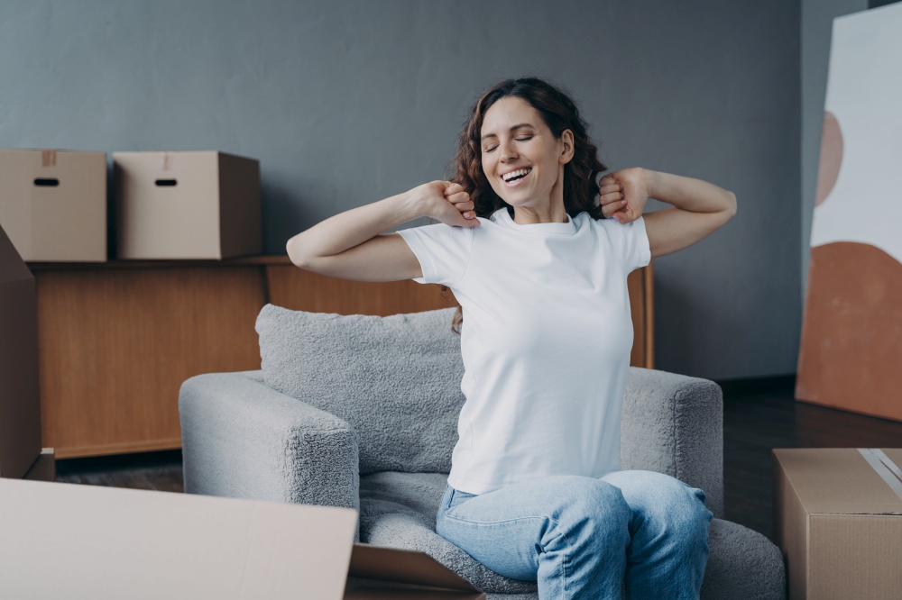 Happy young woman moves alone. Girl is stretching and going to load box. Mover is sitting in armchair among packed cardboard boxes. Independence, life choice and success concept.. Happy young woman moves alone. Girl is stretching and going to load packed box.
