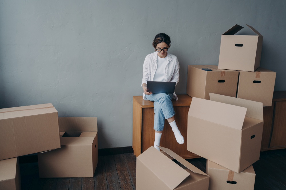 Spanish business lady rents apartment. Young businesswoman moves. Happy girl is sitting among carton boxes and working on laptop. Mortgage loan and real estate purchase.. Spanish business lady rents apartment. Happy girl is sitting among boxes and working on laptop.