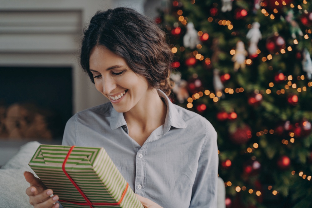 Happy pleased hispanic woman holding christmas gift box while sitting on sofa near xmas tree, celebrating winter holidays at home, smiling european female looking at wrapped New Year present. Happy pleased hispanic woman holding christmas gift box while sitting on sofa near xmas tree