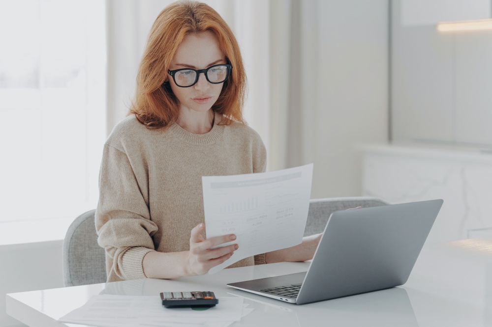 Young focused red-haired woman in spectacles calculating budget at home, sitting at table with laptop, holding paper taxes bills and reading financial information, female managing family budget. Young focused red-haired woman sitting at table with laptop and holding paper taxes bills