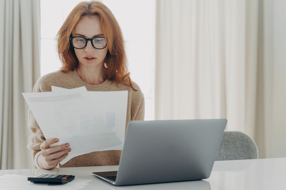Serious redhead Caucasian woman calculates bills holds paper documents has focused look uses laptop computer and calculator thinks how to save money poses at workplace against home interior.. Serious redhead Caucasian woman calculates bills holds paper documents has focused look