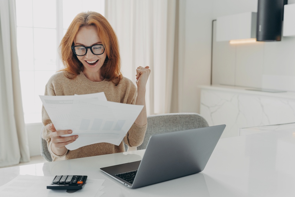 Successful ginger female freelancer checks calculations clenches fist looks happily at paper documents wears spectacles and jumper surrounded by laptop computer and calculator calculates finances. Female freelancer checks calculations clenches fist looks happily at paper documents