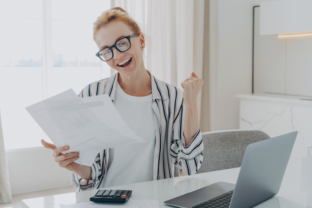Overjoyed excited redhead woman in spectacles exclaiming yes with happy face expression while calculating domestic expenses at home, celebrating money refund or last last mortgage payment. Overjoyed redhead woman exclaiming yes, celebrating money refund or last last mortgage payment