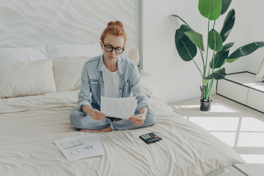 Serious good looking redhead European woman calculates expenses manages household budget looks attentively at paper documents wears denim casual clothes sits on bed against cozy home interior. Good looking redhead European woman calculates expenses manages household budget