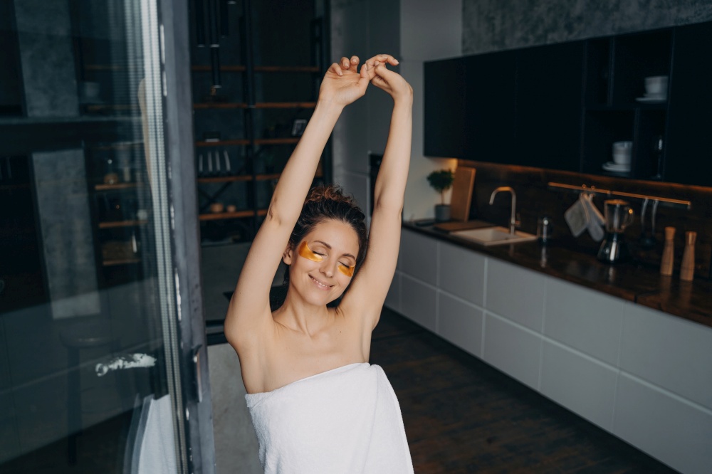 Relaxed woman with under-eye patches stretches in kitchen after shower. Happy girl enjoys morning beauty routine for healthy skin
