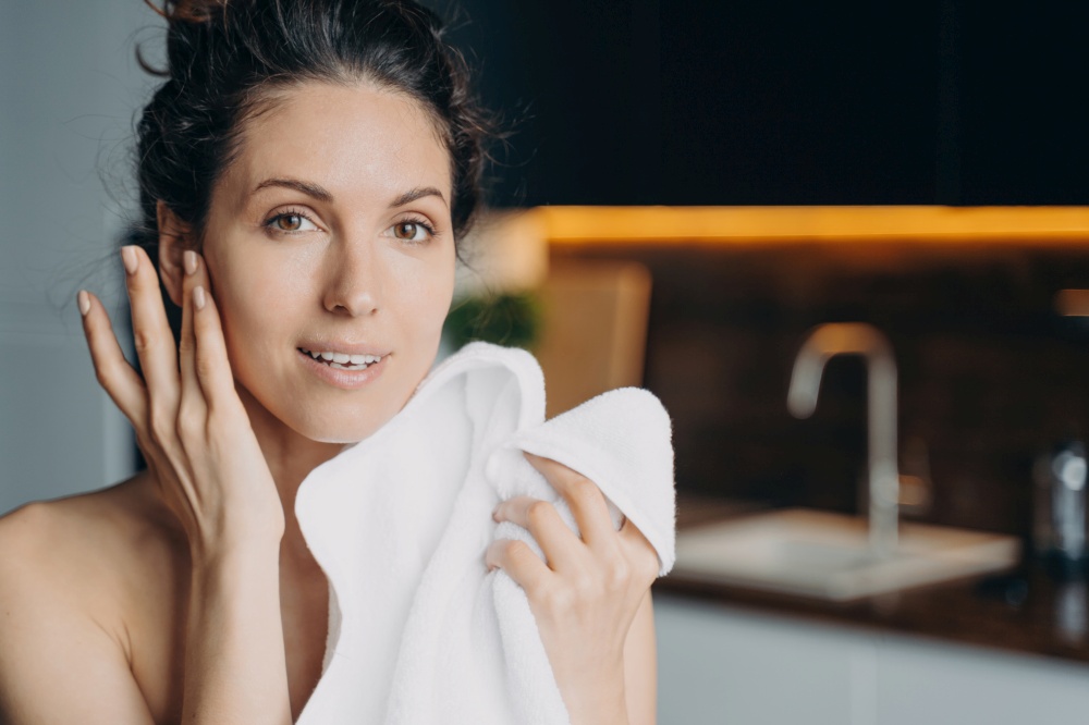 Beautiful woman wipes face with soft towel at home. Spanish female enjoys perfect smooth skin after skincare treatment. Hygiene, self-care concept