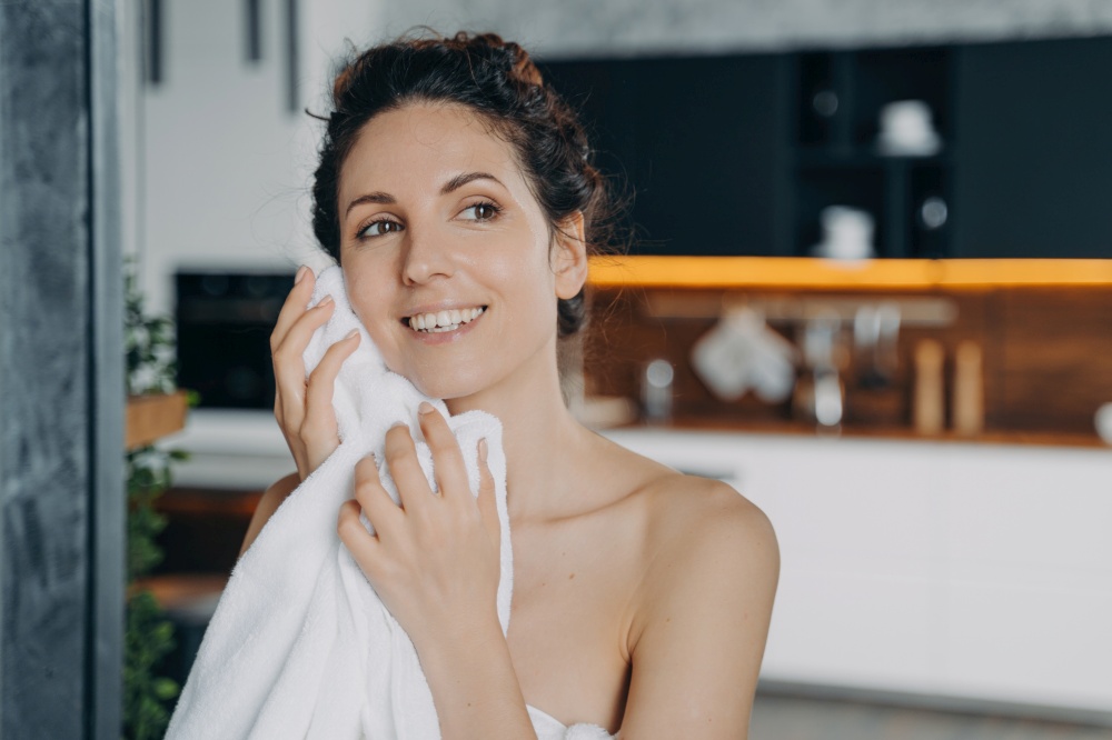 Hispanic woman wipes face with towel at home. Smiling female cleanses, dries facial skin after morning shower. Daily skincare routine.