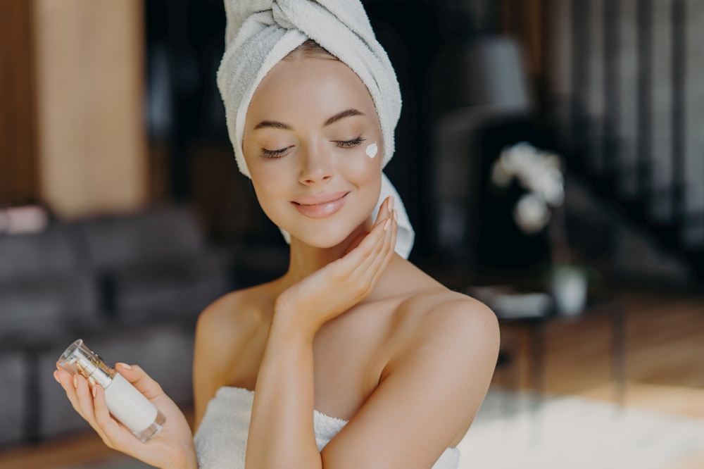 Satisfied woman applies face lotion, wrapped in towel, showcases natural beauty. Cosmetology concept.