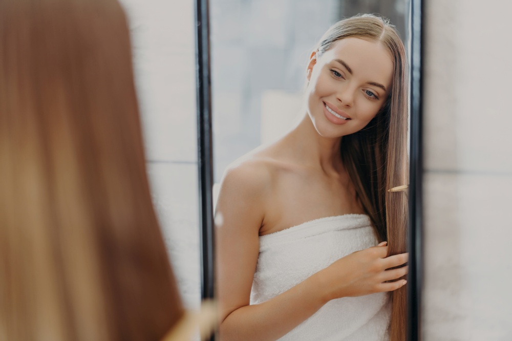 Woman brushes long hair, wrapped in towel, in bathroom. Hair care and beauty concept.
