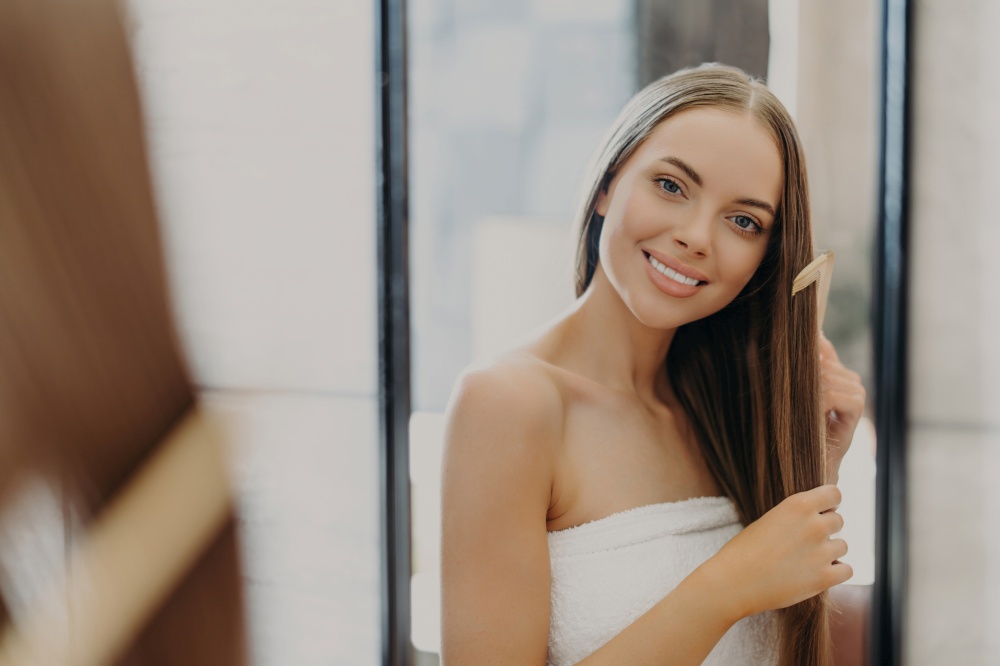 Beautiful woman smiles and combs her straight hair, wrapped in towel, in bathroom. Women&rsquo;s beauty concept