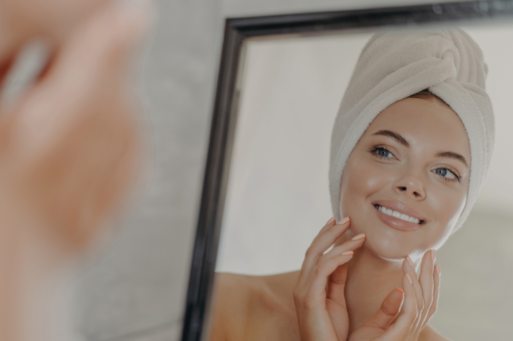 Adorable woman admires reflection, touches healthy skin, towel on head. Beauty routine.