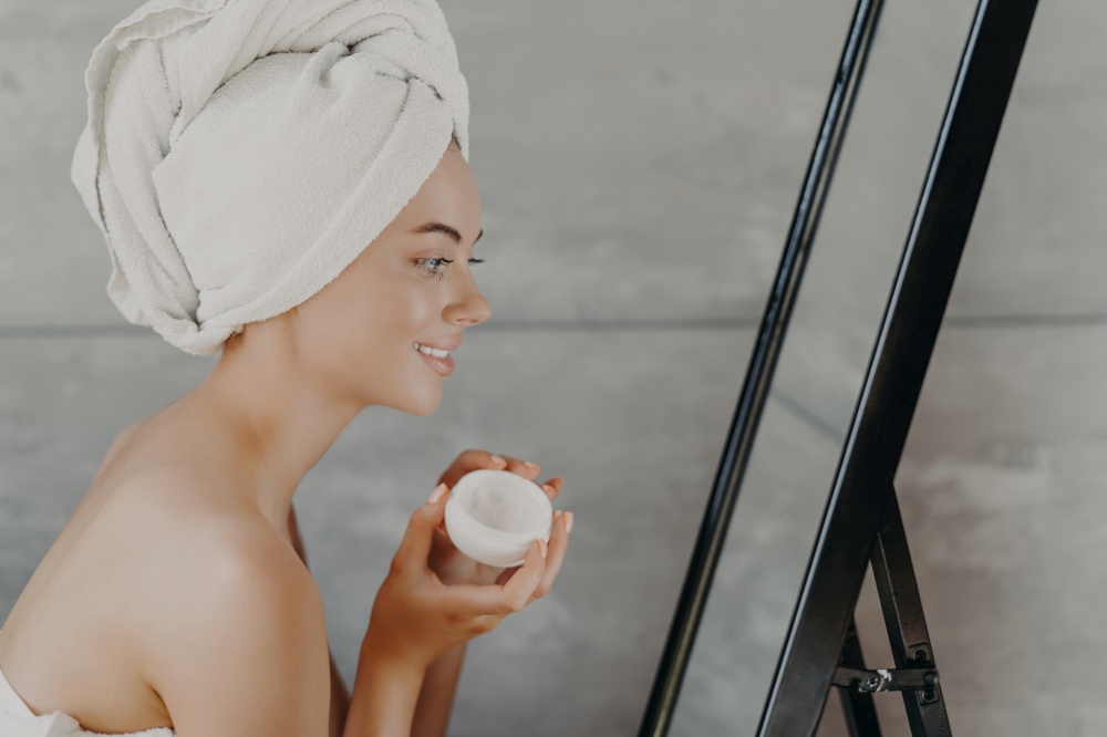 Happy woman with bare shoulders, towel on head, applies face cream, admires reflection. Self-care.