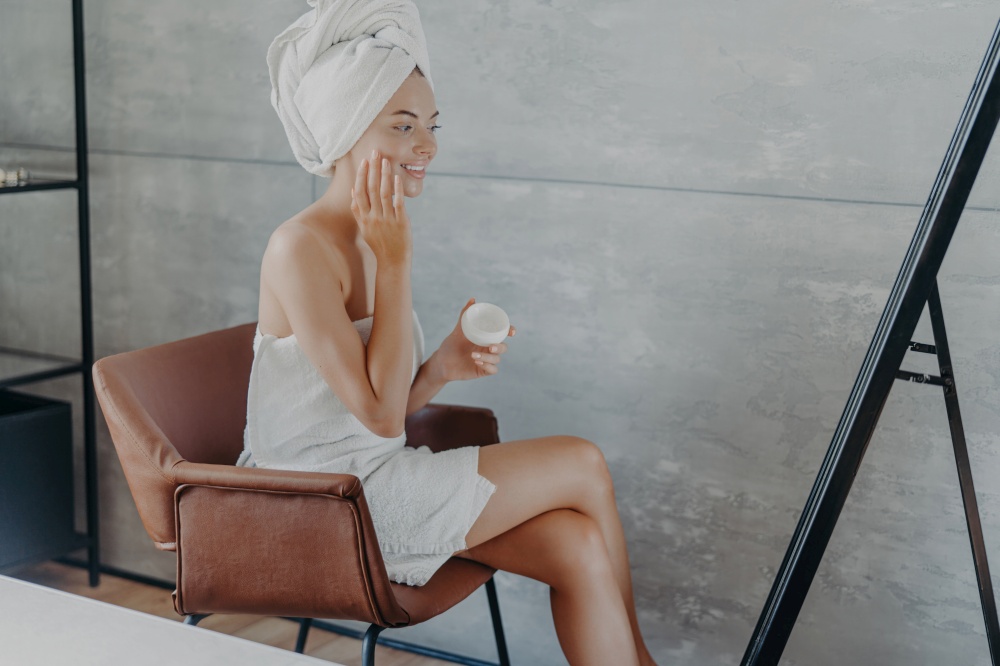 Smiling woman applies face cream, wrapped towel on head, cares for skin. Beauty routine.