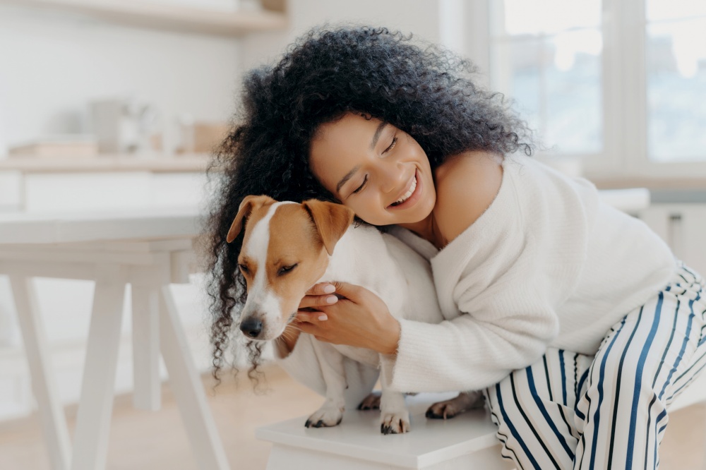 Lovely curly woman embraces beloved dog with care, wearing stylish clothes, posing at home, expressing affection.