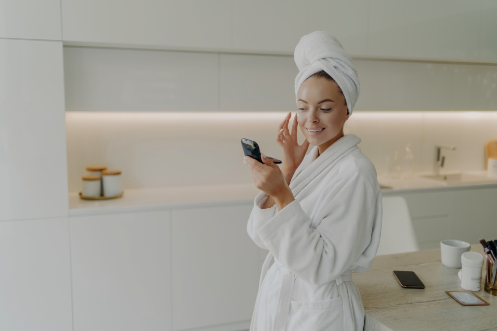 Woman in bathrobe cares for herself, applies skincare product with a smile, uses compact mirror in kitchen.