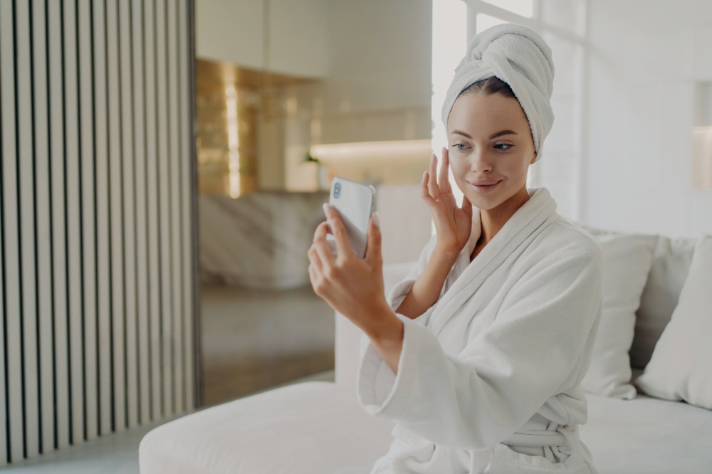 Woman in bathrobe applies cream, touches soft skin, sits on sofa, holds smartphone, beauty routine.