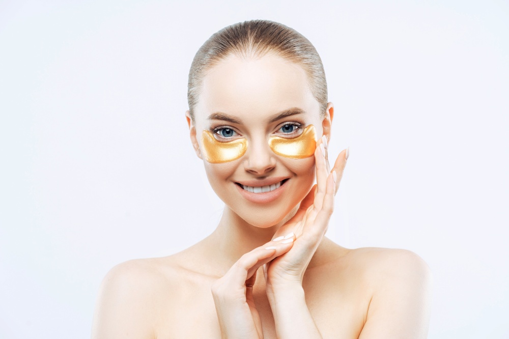 Attractive woman touches face, smiles, applies golden hydrogel eye patches, isolated on white background. Eye mask treatment