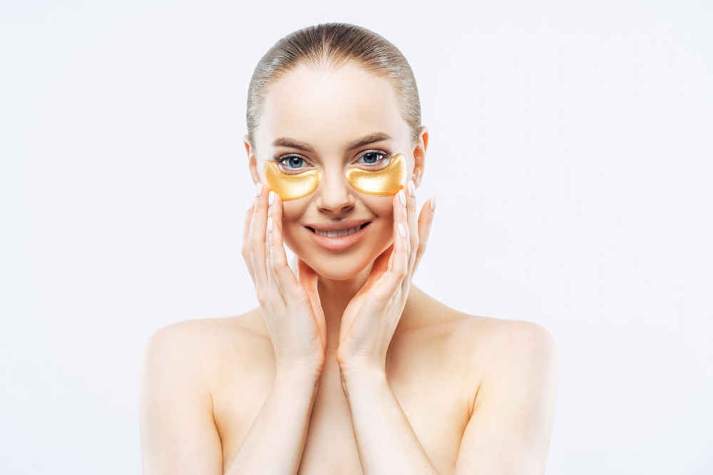Tender European woman smiles, applies under-eye patches, collagen mask for fresh skin, topless against white background