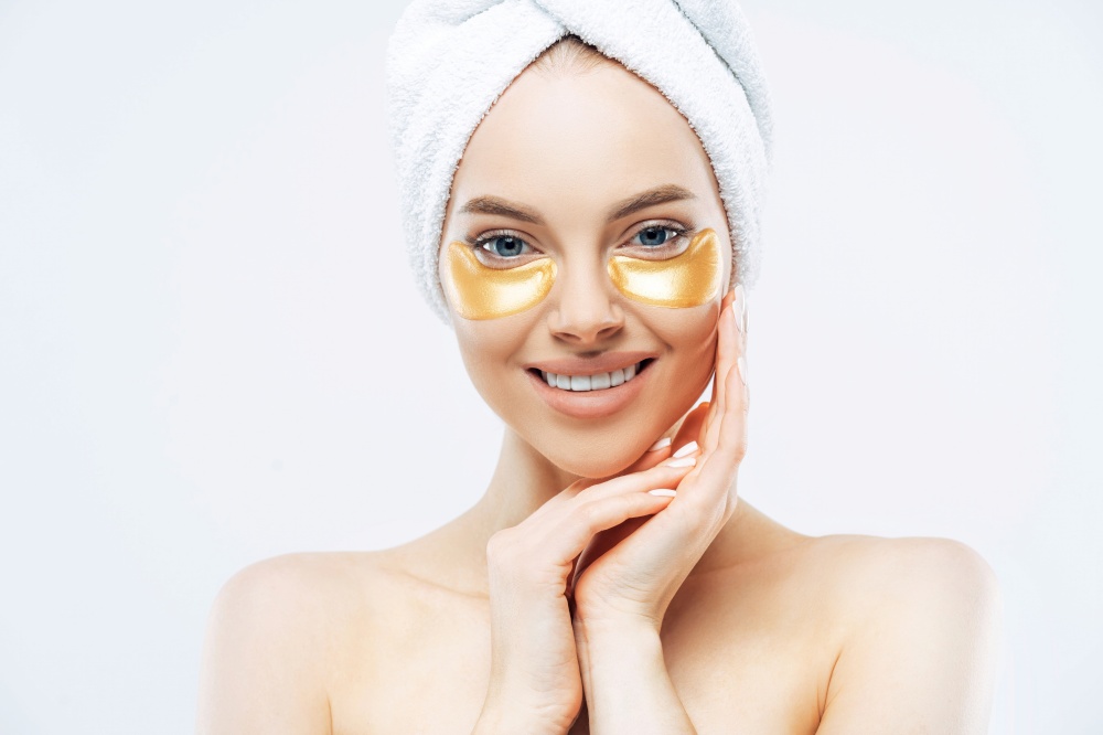 Pretty lady, towel-wrapped head, touches cheeks, applies golden hydrogel patches, shirtless indoors. Removes wrinkles, healthy skin
