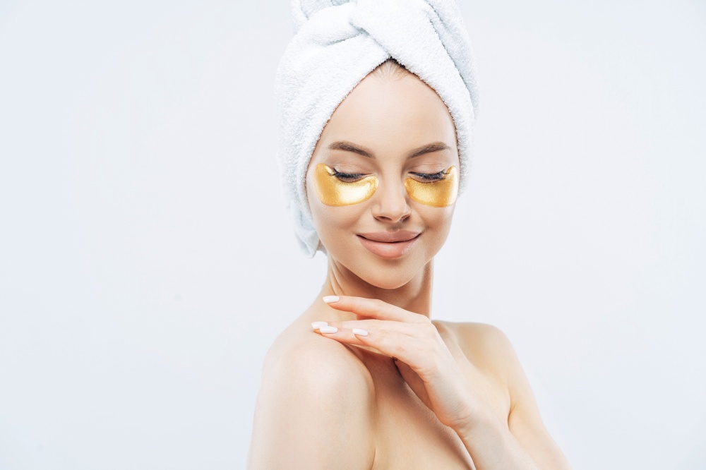 Young woman with gold collagen pads, fresh skin, anti-aging mask, towel on head, touching shoulder.