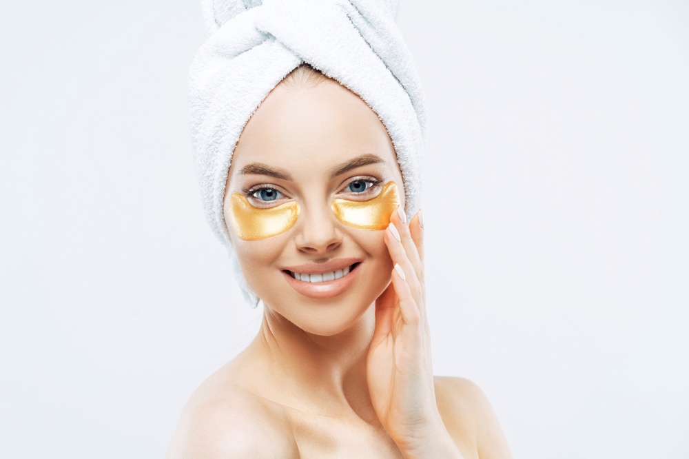 Pretty young woman with golden eye patches, caring for face and skin, towel on head, isolated